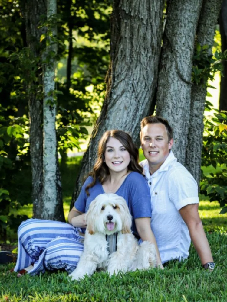 The image features two people sitting on the grass in front of a tree, with a fluffy, light-colored dog in a harness between them. The person on the left wears a blue shirt with striped pants, while the person on the right wears a white shirt.