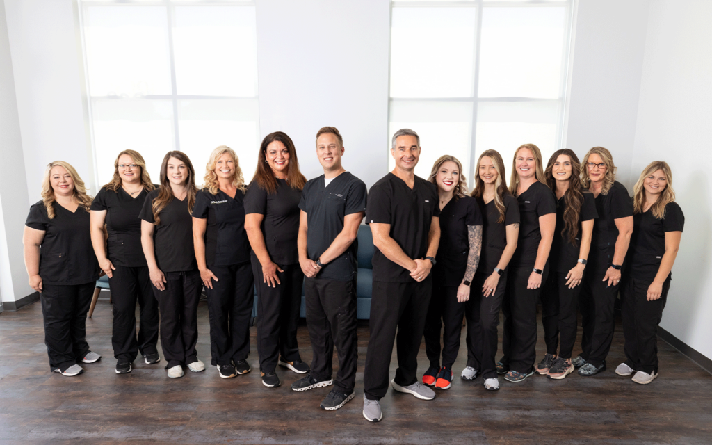 A team of thirteen healthcare professionals in black scrubs stands in a line inside a well-lit room with large windows, wooden floors, and white walls. They are wearing various types of footwear, posing together for a group photo.